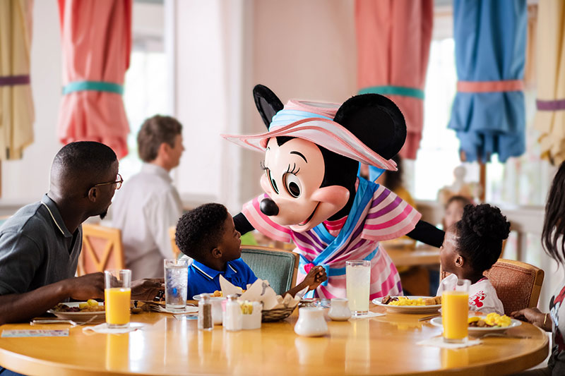 magical-disney-dining-fall-2023-2 Mickey Mouse, dressed as a painter, bends over to entertain a family of four seated for a meal at a restaurant.
